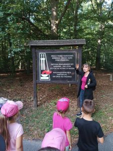 Wychowawczyni oddziały przedszkolnego stoi przy dużej tablicy z napisem "MIEJSCE PAMIĘCI NARODOWEJ BARBARKA - MIEJSCE KAŹNI OFIAR HITLERYZMU" Poniżej napis ten jest powtórzony w języku angielskim i niemieckim. Na przeciwko widać stojące dzieci.