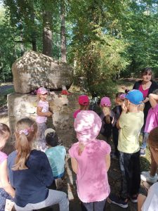 Uczniowie stoją na polanie przy pomniku pamięci. Dzieci zwrócone są plecami. Na przeciwko stoi wychowawczyni oddziału przedszkolnego.