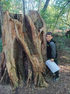 Uczeń przytula się do wielkiego Kawałka pnia drzewa. W ręce trzyma papierową torbę.