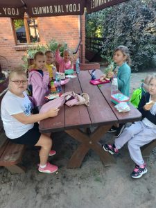 Uczniowie klasy pierwszej siedzą przy dużym drewnianym stole na świeżym powietrzu. Dzieci jedzą kanapki. Na stole stoją rozłożone śniadaniówki.