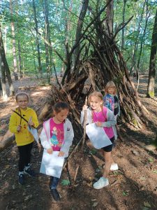Cztery uczennice stoją przy dużym szałasie zbudowanym z gałęzi i patyków. Dziewczynki trzymają papierowe torby. Jedna z nich wspiera się na drewnianym kiju.