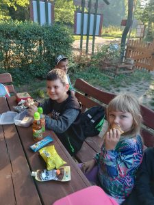 Dwoje uczniów siedzi przy drewnianym stole. Jedzą zabrane przez siebie kanapki. Na stole leżą inne przekąski.