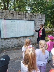 Przy tablicy z napisem "BARBARKA MIEJSCE PAMIĘCI NARODOWEJ" oraz wymienionymi imionami i nazwiskami ofiar, stoi wychowawczyni oddziału przedszkolnego. Wskazuje palcem na tablicę. Na przeciwko widać stojących uczniów.