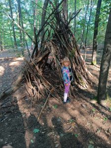 Uczennica stoi przy dużym szałasie zbudowanym z gałęzi i patyków.
