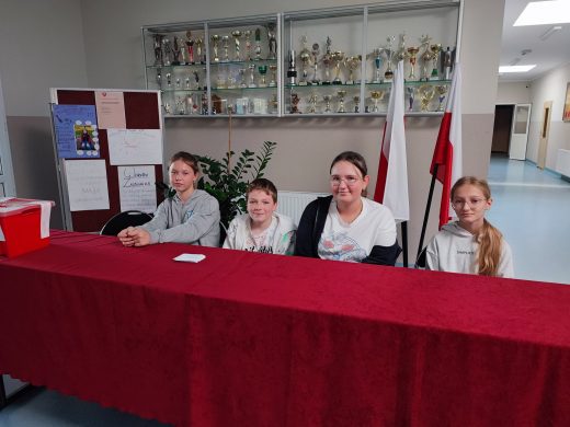 Czworo uczniów wchodzących w skład komisji wyborczej siedzi przy długim stole przykrytym czerwonym materiałem.