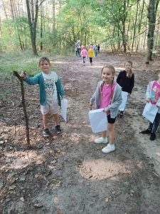 Uczniowie stoją na leśnym szlaku. W dłoniach trzymają papierowe torby. Jeden z uczniów opiera się o drewniana laskę trzymaną w dłoni.