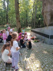 Uczniowie stoją przy pomniku w miejscu pamięci na Barbarce. Część uczniów stoi, część klęczy i się modli. Zdjęcie wykonane bokiem.