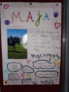 Zdjęcie plakatu ucznia biorącego udział w wyborach do samorządu uczniowskiego. Na białym tle u góry duży kolorowy napis 'Maja". Poniżej zdjęcie dziewczynki na koniu i tekst: "cześć nazywam się Maja Muszyńska Moją pasją są konie chodzę do 3 klasy"; "moje pomysły" i poniżej w chmurkach napisy: "nocka w szkole dla klas 1,3", "dzień piłkarza", "dzień pizzy"; "dzień filmów", "dzień bez plecaka". "głosuj na mnie".