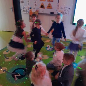 Uczniowie oddziału przedszkolnego ubrani w stroje galowe tańczą na dywanie. Światło jest przygaszone a na dzieciach widać kolorowe światełka. Zdjęcie wykonane w ruchu.