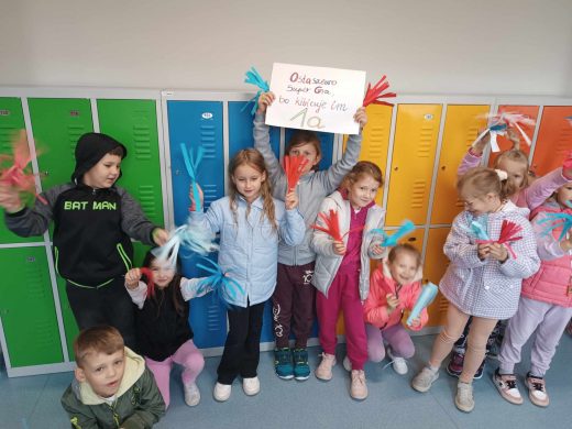 Uczniowie klasy pierwszej stroją przy szkolnych szafkach. Jedna z uczennic trzyma kartkę z kolorowym napisem "Ostaszewo Super Gra bo kibicuje im 1a". Dzieci trzymają w dłoniach wycięte z kolorowych papierów paseczki przypominające małe pompony.