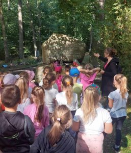 Uczniowie oddziału przedszkolnego stoją przed pomnikiem w kształcie kamienia. Obok nich stoi nauczycielka wskazująca jedną dłonią na pomnik.