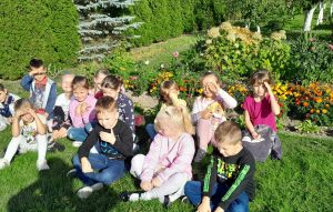 Uczniowie oddziału przedszkolnego siedzą na trawie przed ogródkiem działkowym. dzieci zasłaniają dłońmi oczy przed słońcem.