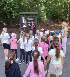 Uczniowie oddziału przedszkolnego stoją na przeciwko tablicy z informacją o miejscu pamięci na Barbarce. Przed tablicą stoi ich nauczycielka podnosząca w górę prawą rękę.