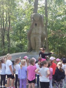 Uczniowie oddziału przedszkolnego stoją przed pomnikiem wyrytym w kamieniu, w kształcie postaci z nachyloną głową i dziurą w miejscu serca. Przed dziećmi stoi Pani nauczycielka wskazując ręką na prawą stronę. Dzieci odwracają się w kierunku przez nią wskazanym.