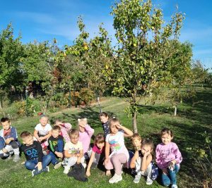Uczniowie oddziału przedszkolnego pozują do wspólnego zdjęcia klęcząc na trawniku przed drzewkami owocowymi. Dzieci zasłaniają dłońmi oczy od słońca.