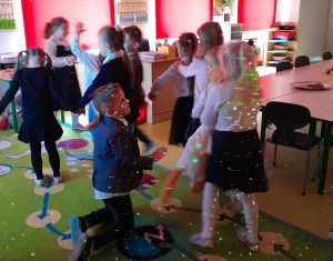 Uczniowie oddziału przedszkolnego ubrani w stroje galowe tańczą na dywanie. Światło jest przygaszone a na dzieciach widać kolorowe światełka. Zdjęcie wykonane w ruchu.