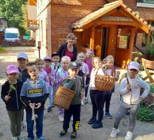 Uczniowie oddziału przedszkolnego stoją wraz ze swoją nauczycielką przed budynkiem z napisem leśniczówka. Dwoje uczniów trzyma w dłoni duże wiklinowe kosze. Część dzieci ma w dłoniach patyki.