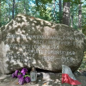 Zdjęcie pomnika w kształcie głazu na którym wyryty jest napis: "ZAMORDOWANYM I ZAMĘCZONYM PRZEZ HITLEROWCÓW NAUCZYCIELOM MIASTA I POWIATU TORUŃSKIEGO HARCERZE MIASTA I POWIATU 1939-1969" . Pod pomnika ustawiony jest znicz, wiązanka sztucznych kwiatów oraz oparta o pomnik polska flaga.