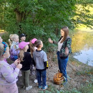 Uczniowie oddziału przedszkolnego stoją na przeciwko Pani, przed której stopami ustawiony jest wiklinowy koszyk. Pani szeroko rozstawia ręce. Za nią widać zejście i staw.