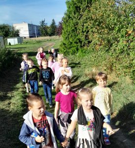 Uczniowie oddziału przedszkolnego idą polną drogą. Po prawej stronie drogi rosną krzewy. Za uczniami widać bloki.