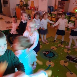 Uczniowie oddziału przedszkolnego ubrani w stroje galowe tańczą na dywanie. Światło jest przygaszone a na dzieciach widać kolorowe światełka. Zdjęcie wykonane w ruchu.