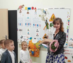 Wychowawczyni stoi z otrzymanymi upominkami od dzieci. Przed nią stoi dwóch uczniów.