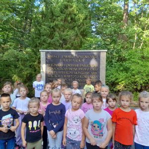 Uczniowie oddziału przedszkolnego pozują do zdjęcia grupowego stojąc przed pomnikiem. na pomniku wyryty jest napis: "MIEJSCE PAMIĘCI NARODOWEJBARBARKA WIEŹNIOM FORTU VII-PATRIOTOM POMORODWANYM PRZEZ OKUPANTA HITLEROWSKIEGO W MIESIĄCACH X XI XII 1839 ROKU SKŁADA HOŁD SPOŁECZEŃSTWO MIASTA I WOJEWÓDZTWA". Część napisu zasłonięta jest przez uczniów. Wszystkie dzieci mają poważne miny.