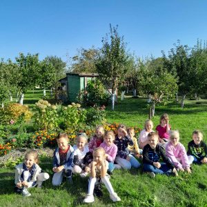 Uczniowie oddziału przedszkolnego siedzą na trawie. Za ich plecami widać ogód z drzewkami i altaną.