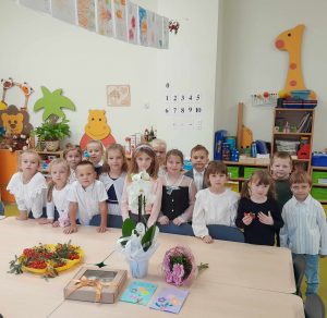 Uczniowie oddziału przedszkolnego ubrani w galowe stroje pozują do wspólnego zdjęcia. Przed dziećmi stoi duży stół , na którym ułożone są upominki dla wychowawczyni - kwiaty, laurki i pudełko z prezentem. W tle widać salę lekcyjną.