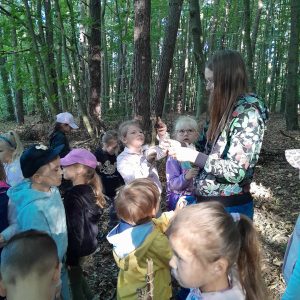 Uczniowie stoją w lesie. Są zgromadzeni wokół stojącej wśród nich Pani. Jedna z uczennic wysoko podnosi w górę trzymaną w dłoni szyszkę.