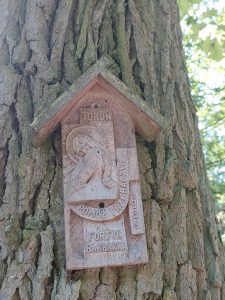 Zdjęcie zamieszczonego na drzewie pomnika w kształcie domku dla ptaków. Na pomniku widać napisy "TORUŃ", POMÓŻ PRZEBACZYĆ", "1939" oraz "FORT VII BARBARKA". Między napisami umieszczony jest wyryty w drewnie święty obrazek przedstawiający Chrystusa tulącego głowę postaci stojącej naprzeciwko.