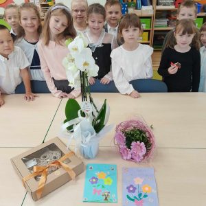 Uczniowie oddziału przedszkolnego ubrani w galowe stroje pozują do wspólnego zdjęcia. Przed dziećmi stoi duży stół , na którym ułożone sa upominki dla wychowawczyni - kwiaty, laurki i pudełko z prezentem.