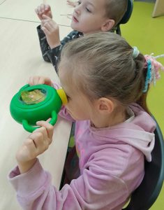 Uczennica trzyma w dłoniach zielone urządzenie, które przykłada do jednego oka. Dzięki urządzeniu widzi w powiększeniu umieszczony w nim liść drzewa. Dziewczynka siedzi przy stoliku a obok niej widać siedzącego innego ucznia.