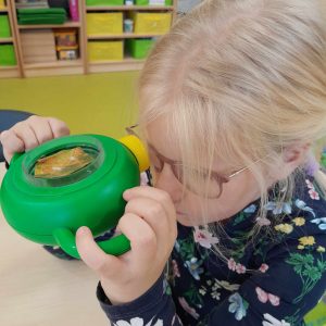 Uczennica trzyma w dłoniach zielone urządzenie, które przykłada do jednego oka. Dzięki urządzeniu widzi w powiększeniu umieszczony w nim liść drzewa.