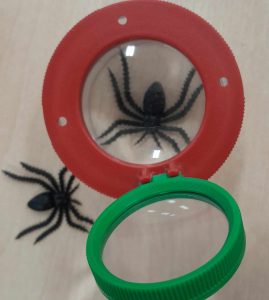 Na stoliku leżą ułożone dwa plastikowe pająki. Jeden z nich jest widoczny w powiększeniu przez lupę ustawioną nad nim. Szkła umieszczono w dwóch plastikowych kółkach - czerwonym i zielonym. nad pająkiem ustawione jest szkło w czerwonej oprawie do którego przyczepiono jest szkoło w zielonym obramowaniu.