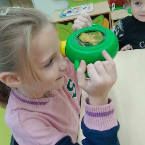 Uczennica trzyma w dłoniach zielone urządzenie, które przykłada do jednego oka. Dzięki urządzeniu widzi w powiększeniu umieszczony w nim liść drzewa.