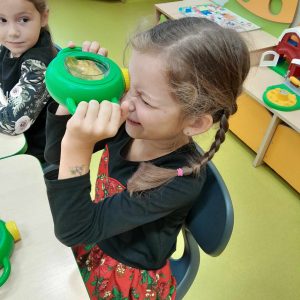 Uczennica trzyma w dłoniach zielone urządzenie, które przykłada do jednego oka. Dzięki urządzeniu widzi w powiększeniu umieszczony w nim liść drzewa. Dziewczynka siedzi przy stoliku a obok widać patrząca na nią koleżankę siedzącą obok.