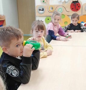 Przy długim stole siedzi czworo uczniów. Jeden z uczniów trzyma przed okiem urządzenie służące do powiększania przedmiotów. Pozostałe uczennice patrzą na chłopca.