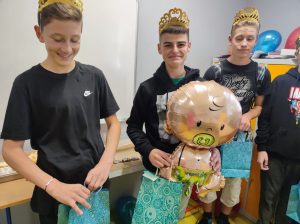 Trzech uczniów klasy siódmej stoi obok siebie z koronami na głowach. uczeń stojący po środku trzyma balona w kształcie dzidziusia ze smoczkiem w buzi.