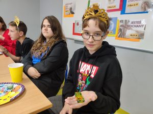 Uczeń klasy siódmej siedzi przy stole z koroną na głowie. Chłopiec trzyma w wyciągniętej dłoni samochodzik. Obok siedzi uczennica spoglądająca w jego stronę.