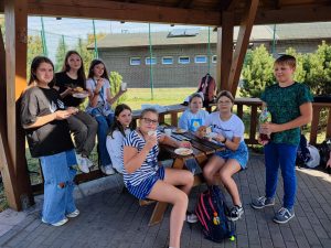 Uczniowie klasy szóstej siedzą przy ławeczce pod zadaszeniem. Dzieci trzymają kubki z piciem i talerzyki papierowe.