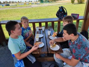 Uczniowie klasy szóstej siedzą przy drewnianym stole pod zadaszeniem. Chłopcy jedzą pizze. Na stole leżą papierowe kubki i talerzyki.