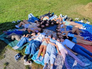 Uczniowie klasy szóstej leżą na ułożonych na trawniku kolorowych kocach. dzieci stykają się głowami tworząc okrąg.
