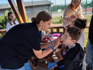 Uczennica klasy szóstej pochyla się nad swoim kolegą by pomalować mu twarz. chłopiec ma zamknięte oczy. W tle widać innych uczniów.