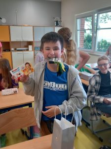 Uczeń klasy piątej stoi przy stoliku w jednej ręce trzyma czekoladki a w zębach skarpetki. W ten sposób prezentuje otrzymany od koleżanek prezent.