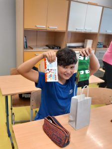 Uczeń klasy piątej trzyma na wysokości uszu otrzymany prezent - z jednej strony czekoladki, z drugiej skarpetki. Chłopiec siedzi za stołem na którym leży piórnik i stoi szara torebka prezentowa.