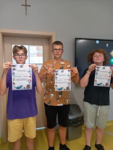 Trzech uczniów klasy ósmej stoi w rzędzie trzymając w palcach przed sobą otrzymane dyplomy.