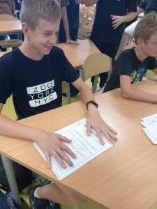 Uczeń klasy ósmej siedzi przy stoliku. Na kartce papieru ma rozłożone dłonie z pomalowanymi na kolorowo paznokciami.