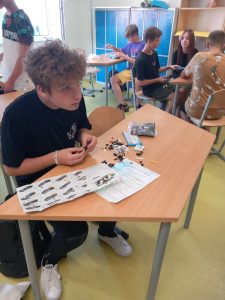 Uczeń klasy ósmej siedzi przy stoliku na którym ma rozłożona instrukcję i klocki lego. Chłopiec składa klocki w dłoniach i patrzy się przed siebie.