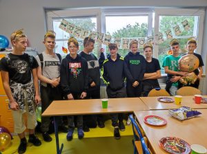 Uczniowie klasy siódmej stoją w rzędzie na tle okna. Za nimi na girlandzie rozwieszony jest napis "DZIEŃ CHŁOPAKA". Chłopcy na głowach mają korony.